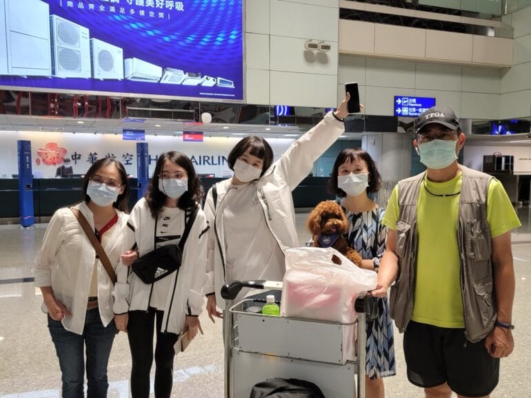 五名戴口罩的人在機場與小狗及行李推車合照,中間的人舉起手機